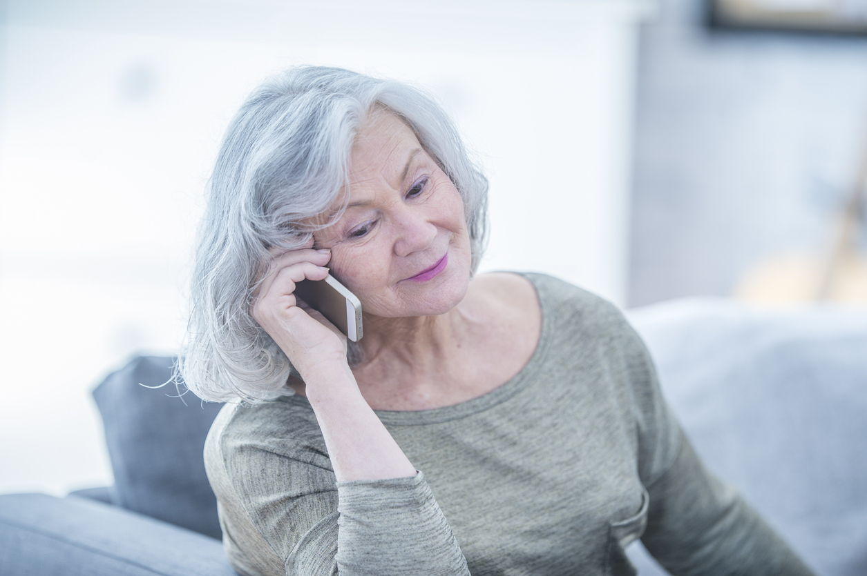 A Caucasian senior woman with white hair is indoors in her house. She is sitting on the sofa and talking on her smartphone.