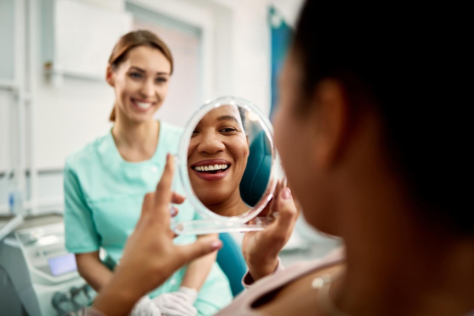 Close up of African American woman using mirror while looking at her teeth after dental check up at dentist's.